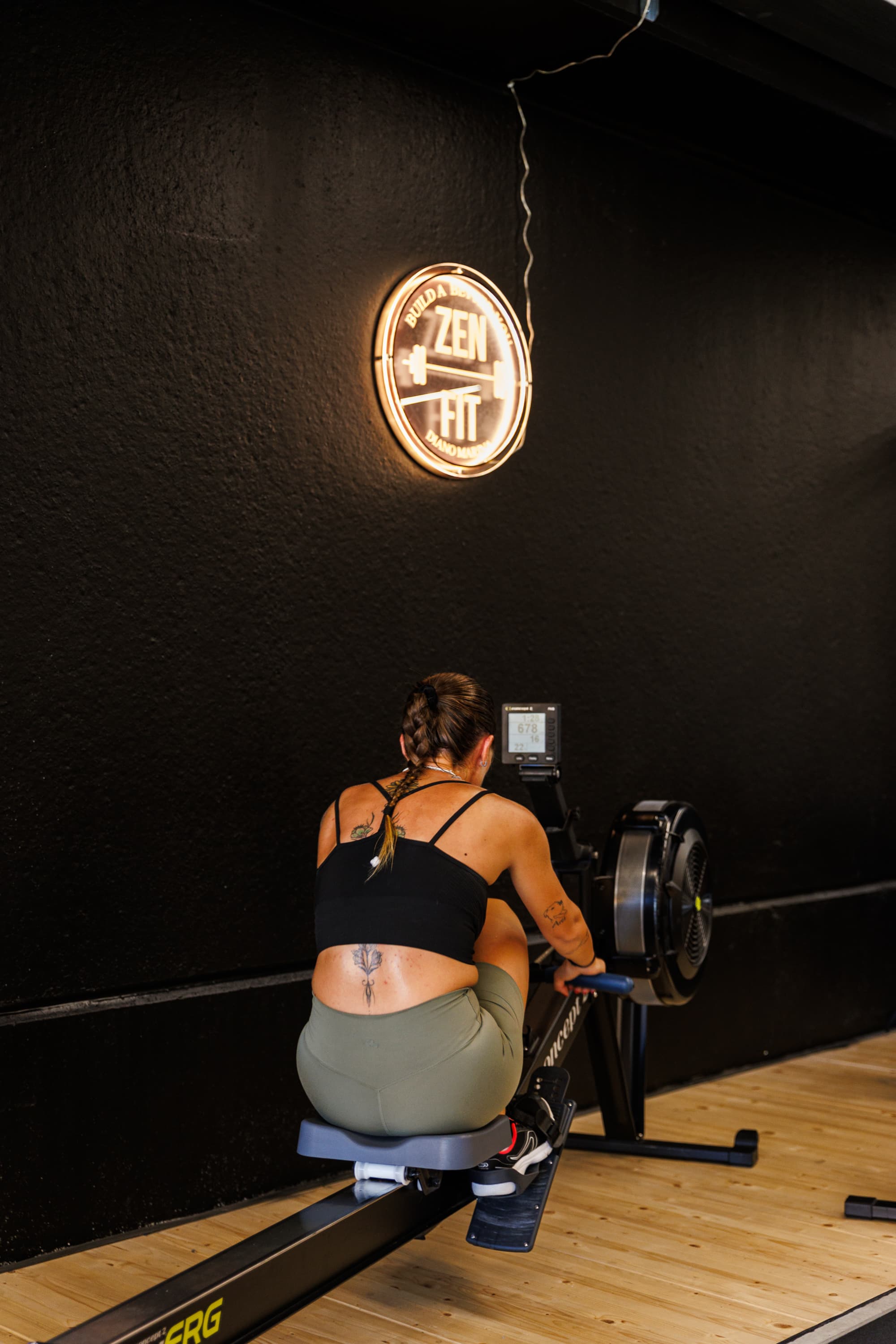 Una ragazza al vogatore contro la parete nera, sotto l'insegna luminosa ZENFIT.