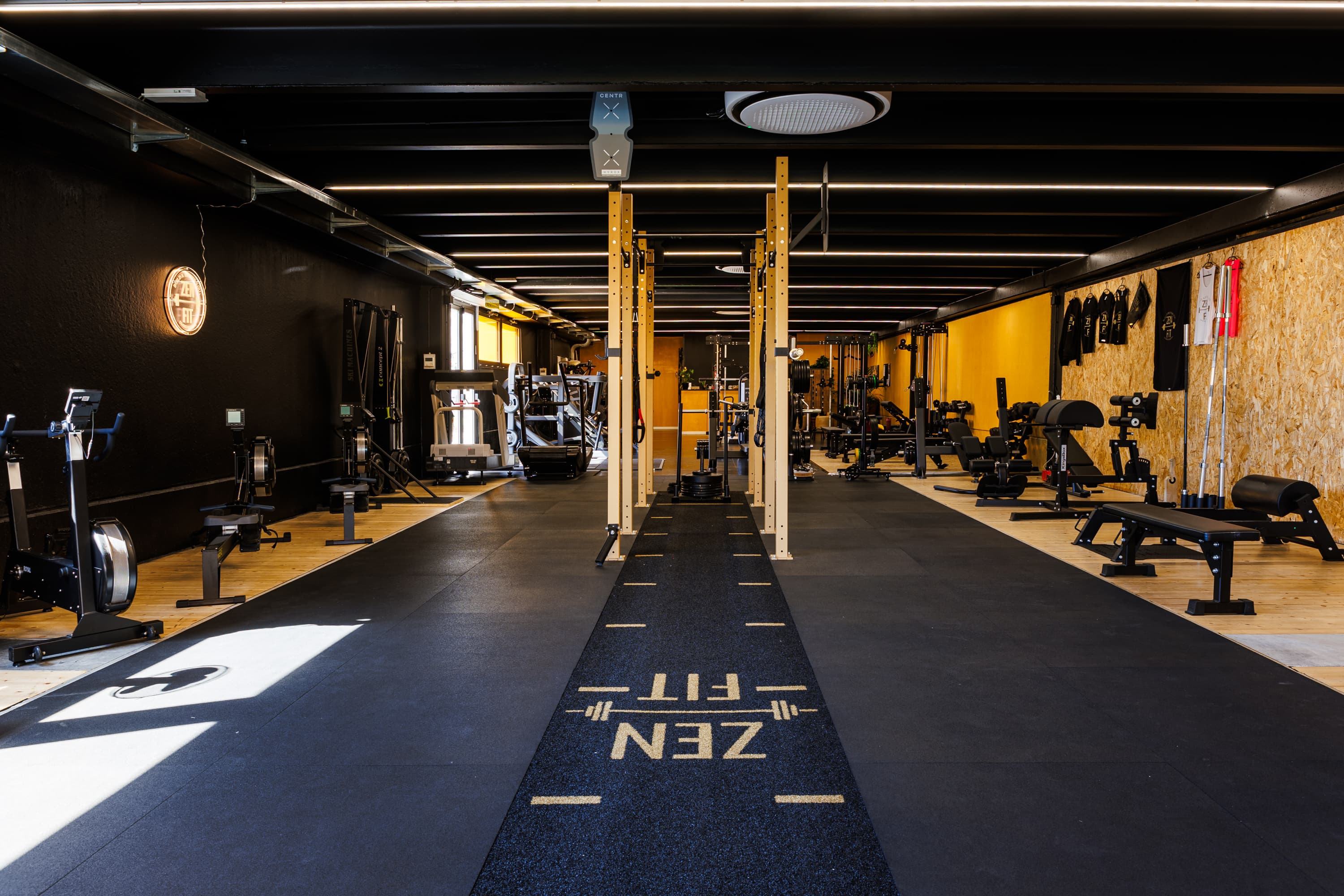 La corsia dell'area functional con la scritta ZENFIT sul pavimento, fra i rack in legno e la parete gialla.